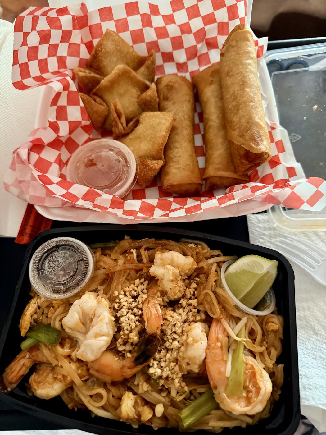 A basket of pork eggrolls above a takeout box of shrimp pad Thai with lime and crushed peanuts